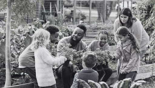 A multi-generational group working together in a community garden, including children holding fresh produce they helped harvest.