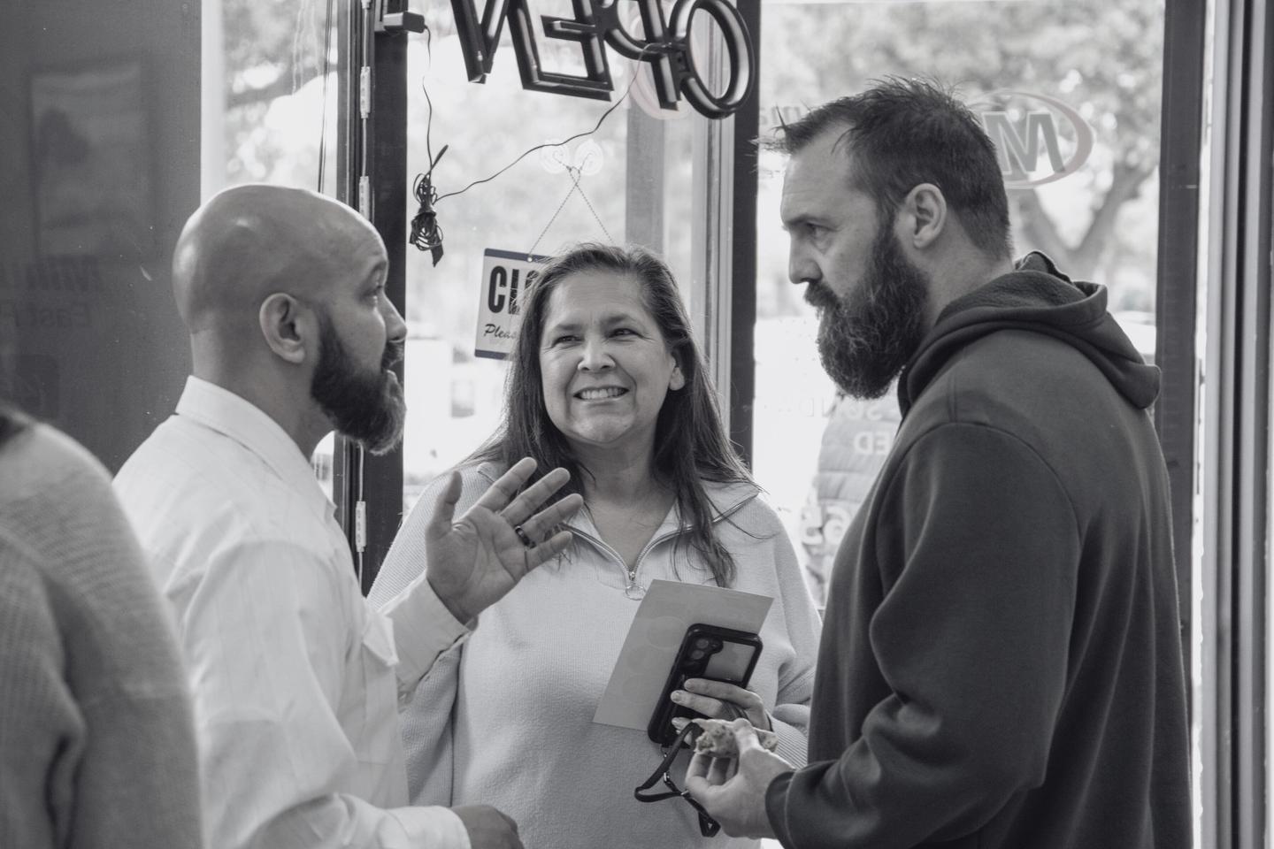 Three business owners in focused conversation outside a storefront, one gesturing as the other two listen.